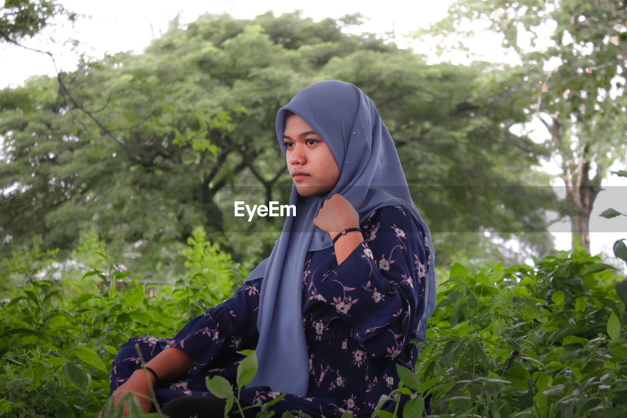 Young woman looking away while standing on land against trees