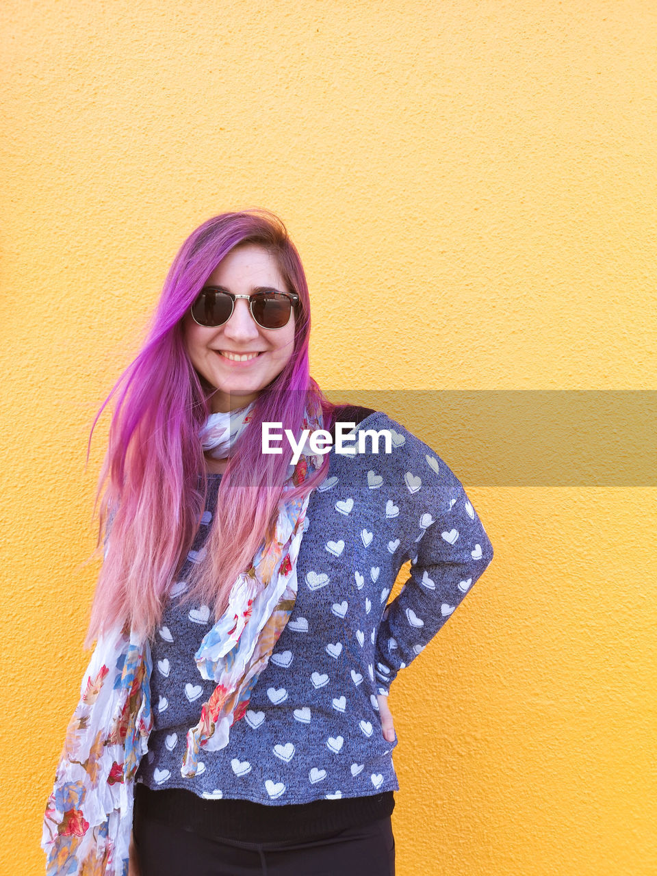 Portrait of smiling young woman with bright purple hair standing against yellow wall