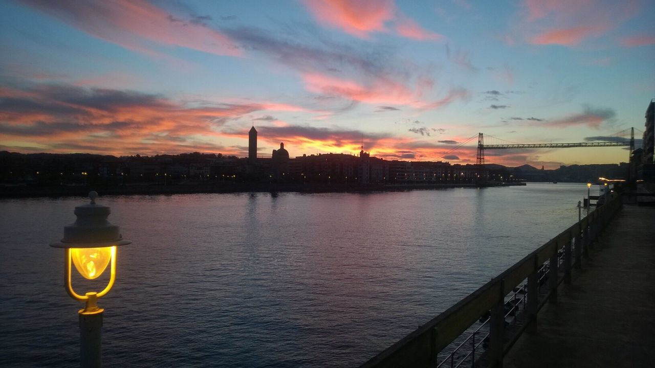 RIVER BY ILLUMINATED CITY AGAINST SKY DURING SUNSET