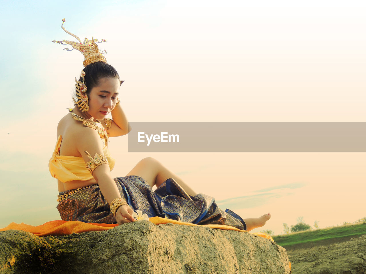 Full length of woman in traditional clothing sitting on rock against sky during sunset