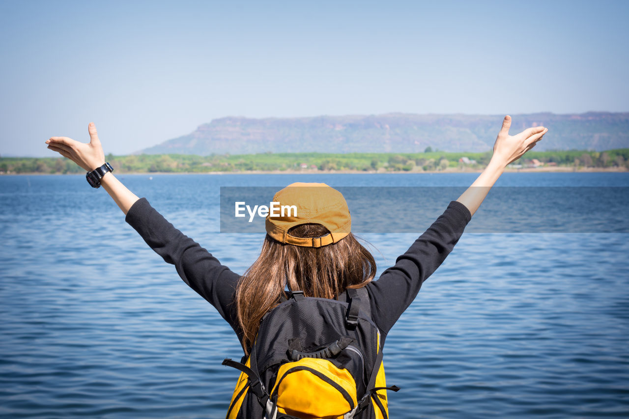 Rear view of backpack woman with arms raised standing by lake against clear sky