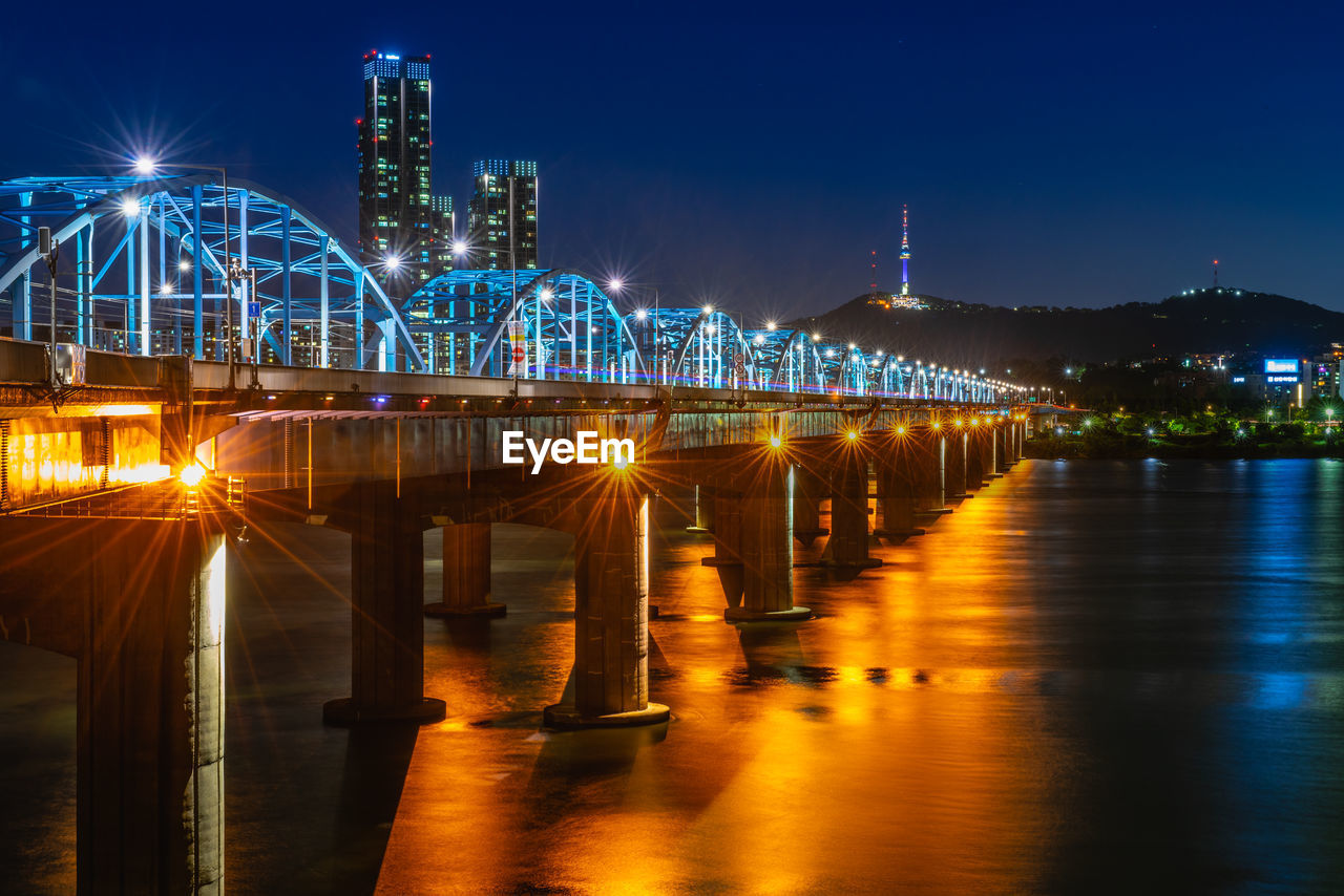 Illuminated bridge over river by buildings against sky at night