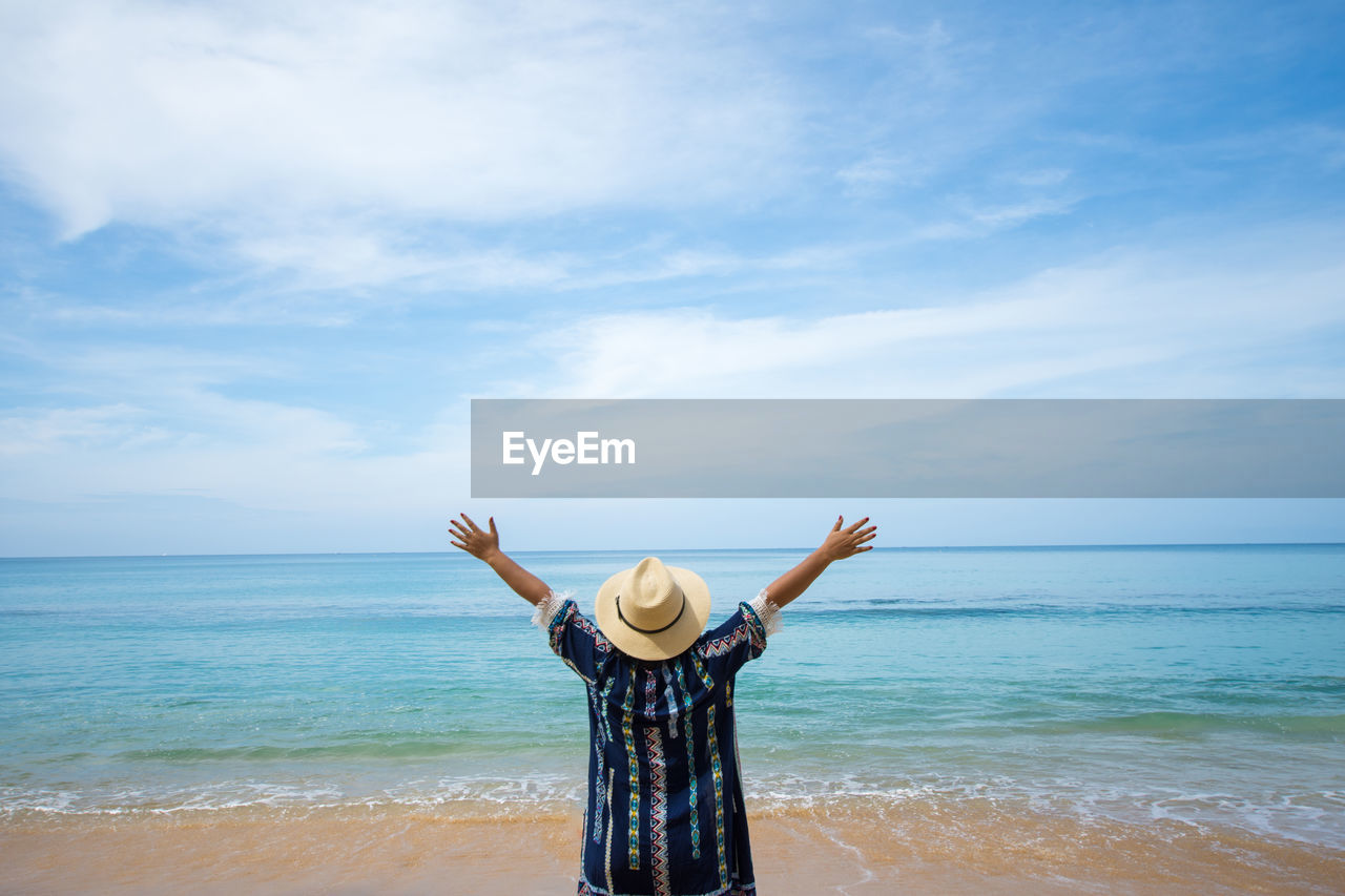 Rear view of carefree woman with arms raised standing at beach against sky