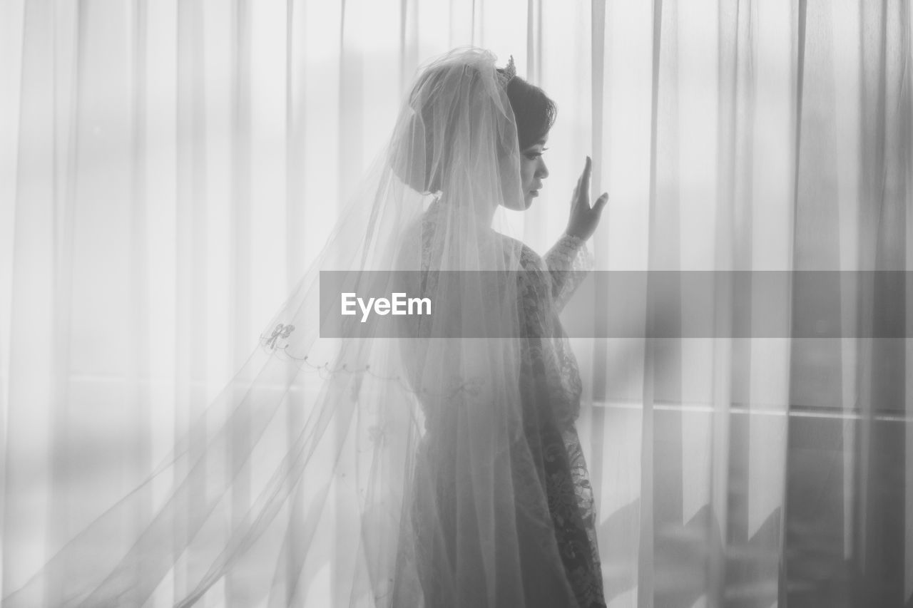 Young woman wearing wedding dress standing by window at home