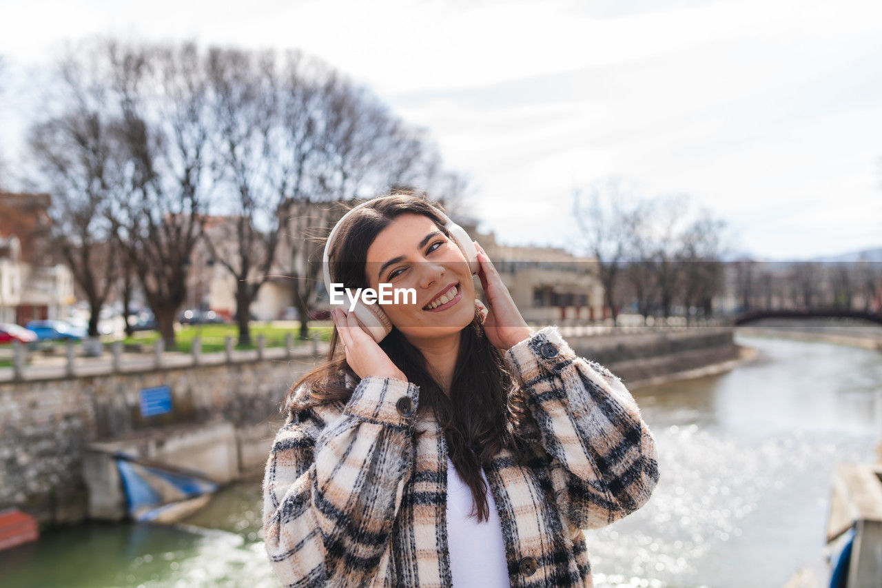 one person, smiling, adult, women, happiness, water, young adult, nature, emotion, spring, portrait, winter, clothing, leisure activity, lifestyles, enjoyment, teeth, warm clothing, smile, architecture, cheerful, long hair, day, hairstyle, waist up, looking, sky, positive emotion, outdoors, human face, brown hair, tree, casual clothing, female, person, front view, standing, holiday, cold temperature, river, copy space, travel destinations, city, technology, carefree, photo shoot, mobile phone, looking away, vacation, tourism, travel, trip, bare tree, smartphone