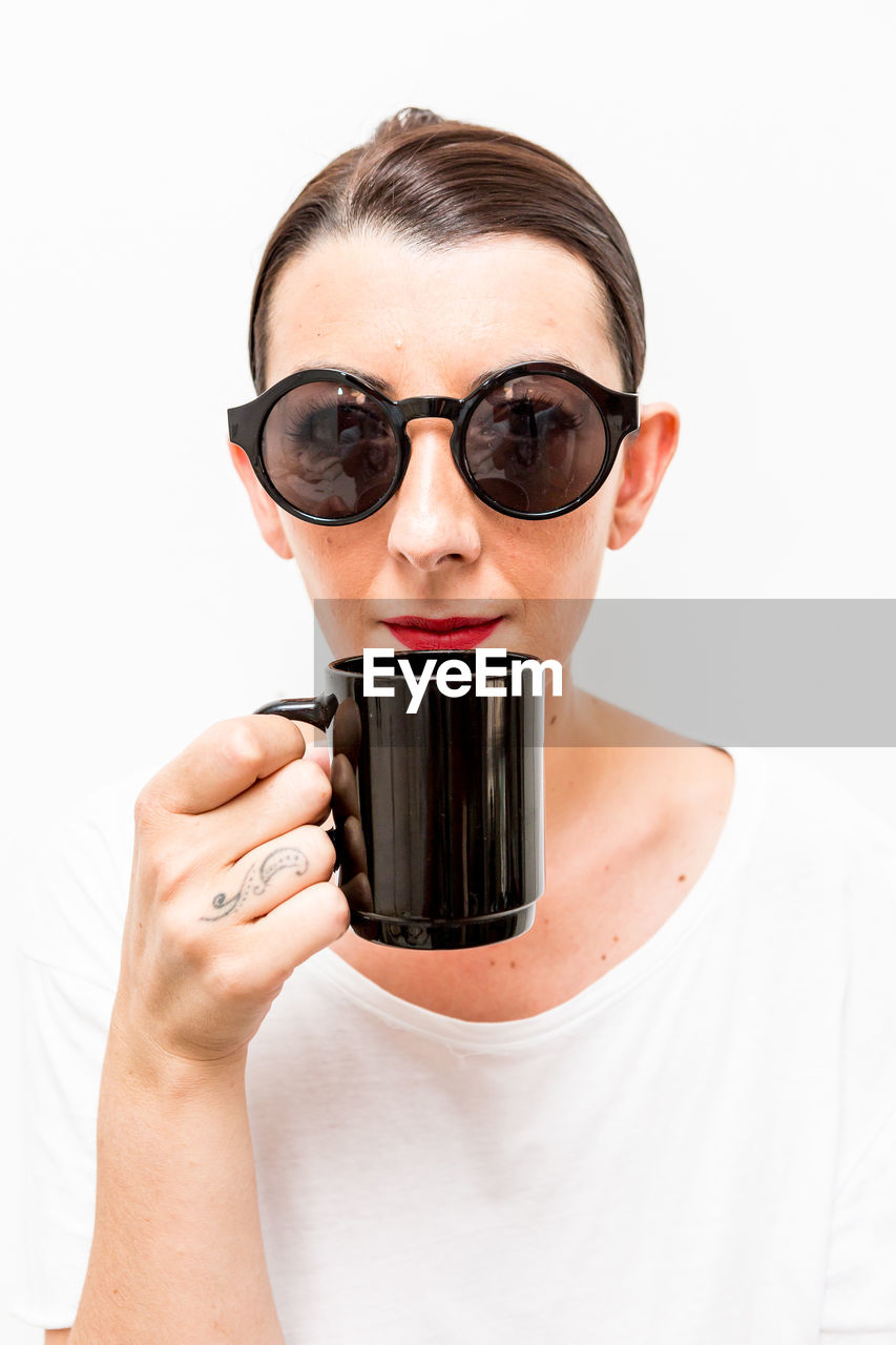 Portrait of young woman wearing sunglasses having coffee against white background