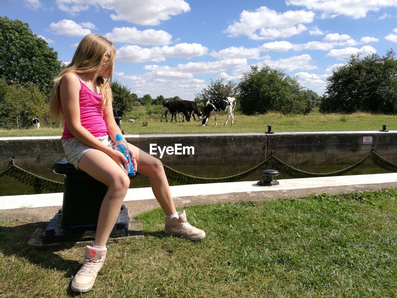 Girl sitting on bollard while looking at horses on grassy field against cloudy sky