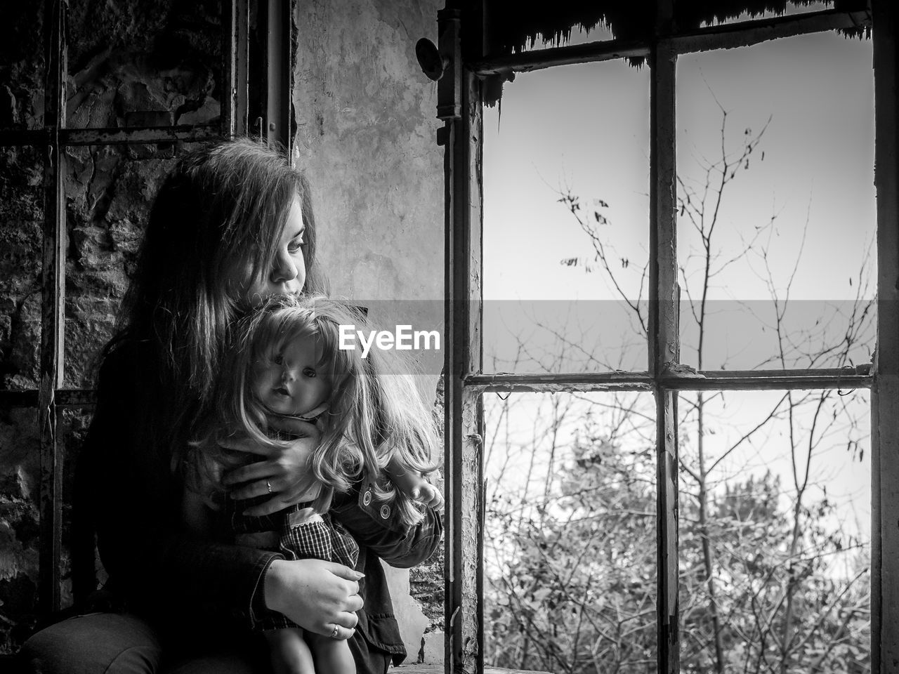 Young adult sitting and holding a doll on an abandoned room near a window.