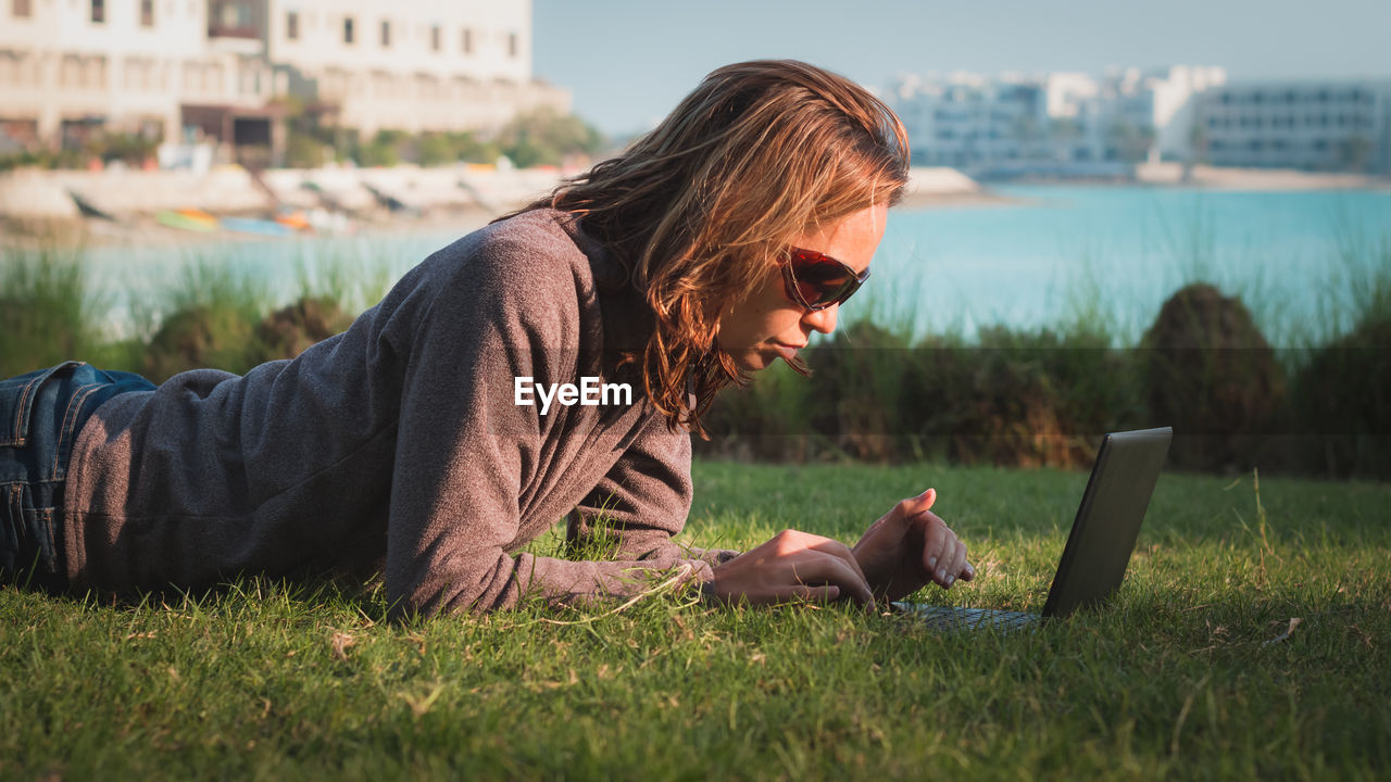 Woman working on laptop while lying on front at grassy field against lake