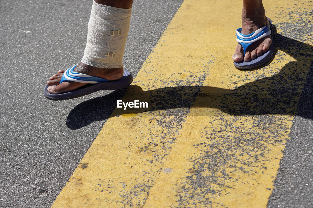 low section, yellow, human leg, shoe, shadow, road, limb, human limb, day, lifestyles, sunlight, one person, city, street, high angle view, road marking, standing, symbol, men, leisure activity, marking, blue, adult, outdoors, transportation, asphalt, sign, footwear, nature, spring, human foot, casual clothing, sneakers, road surface, footpath, women