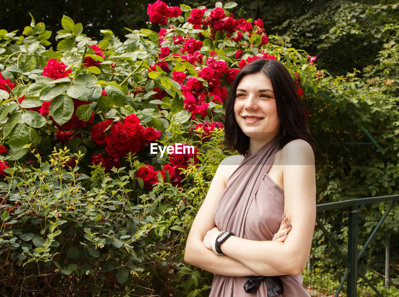 PORTRAIT OF A SMILING YOUNG WOMAN STANDING BY PLANTS