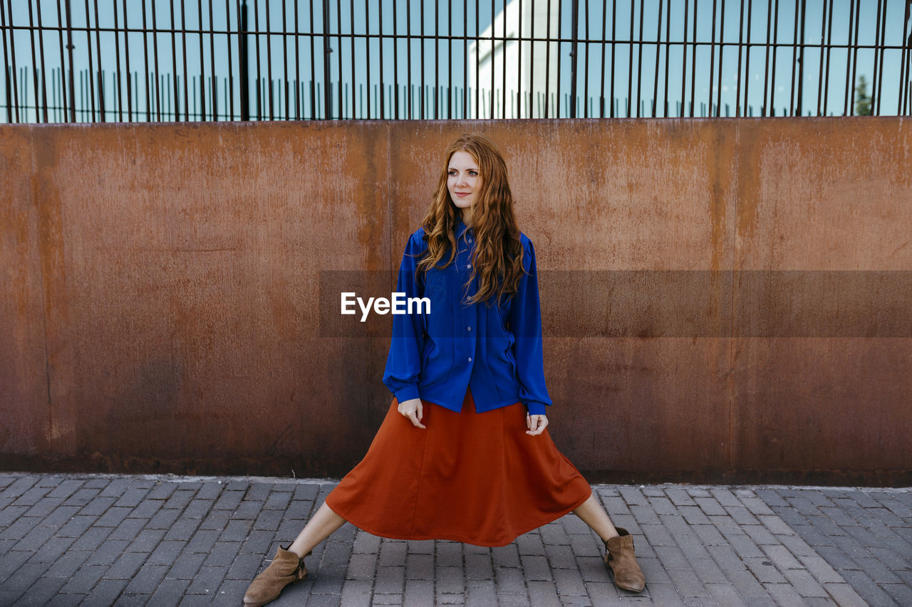 Young woman standing with legs apart on footpath against brown wall