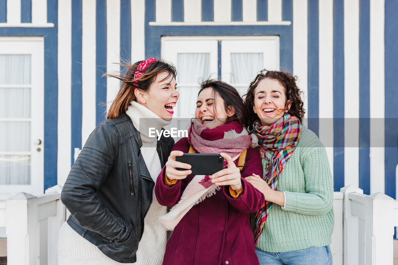 Happy women friends using mobile phone in front of colorful houses.costa nova, aveiro, portugal