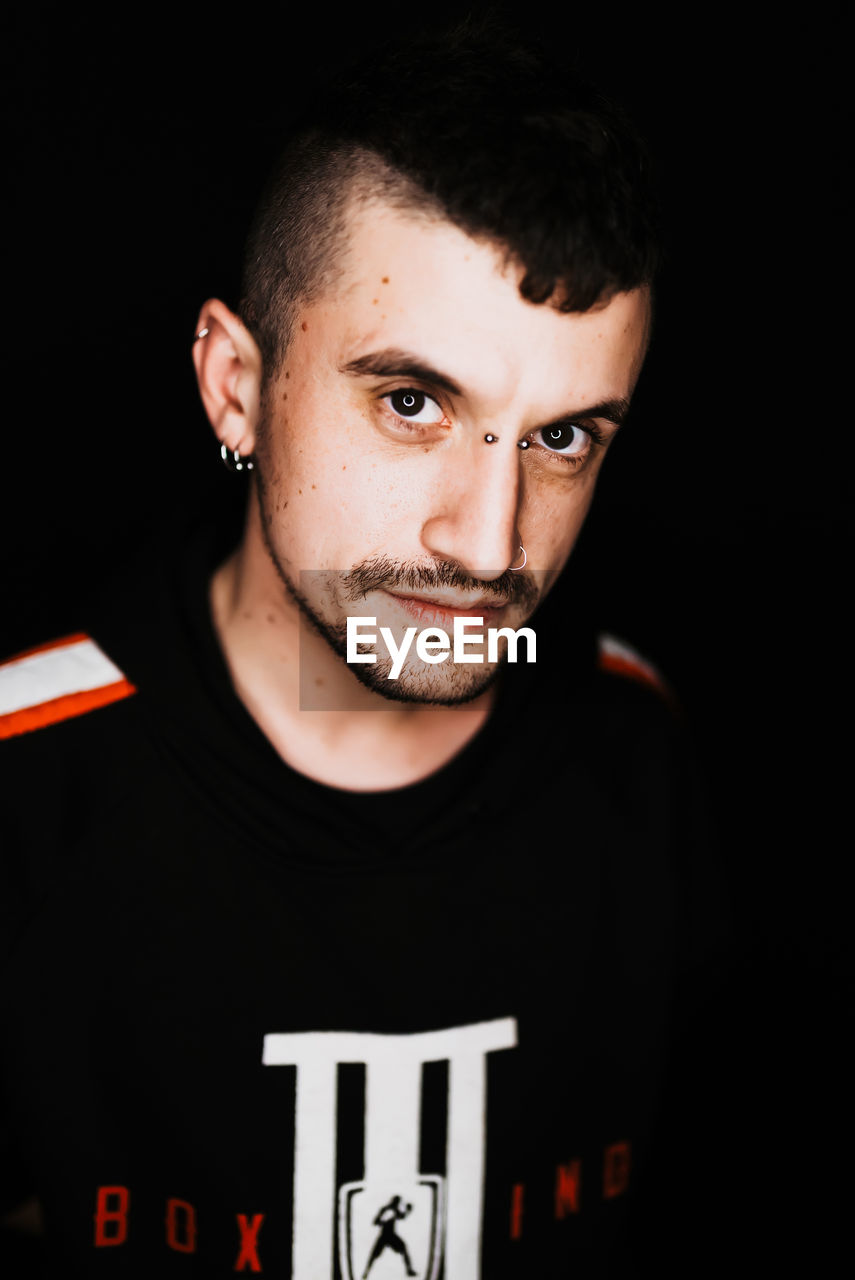 Confident adult male with piercing and mohawk smiling and looking at camera in dark room