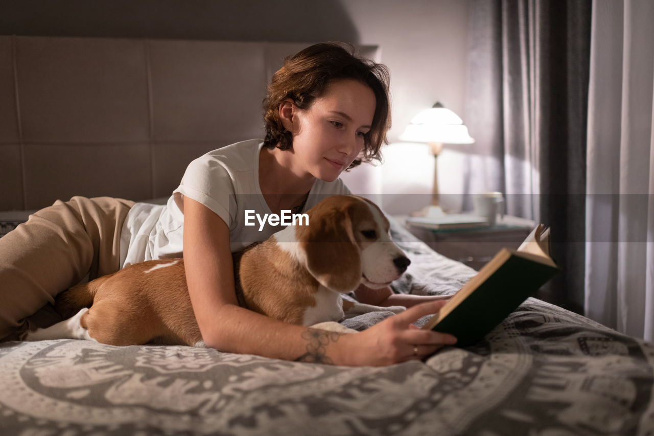 Young woman with beagle reading book on bed