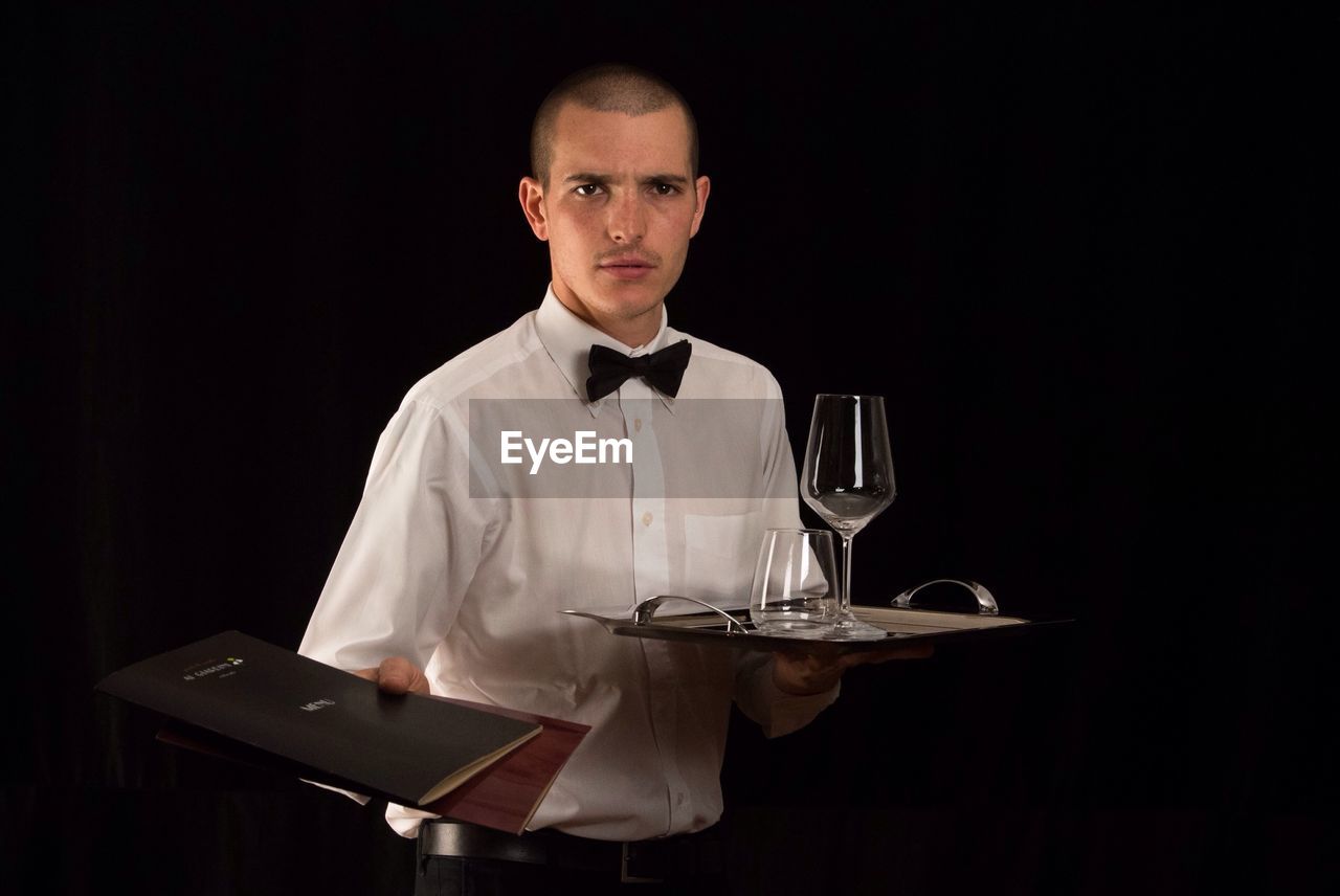 Portrait of waiter holding tray while offering menu over black background