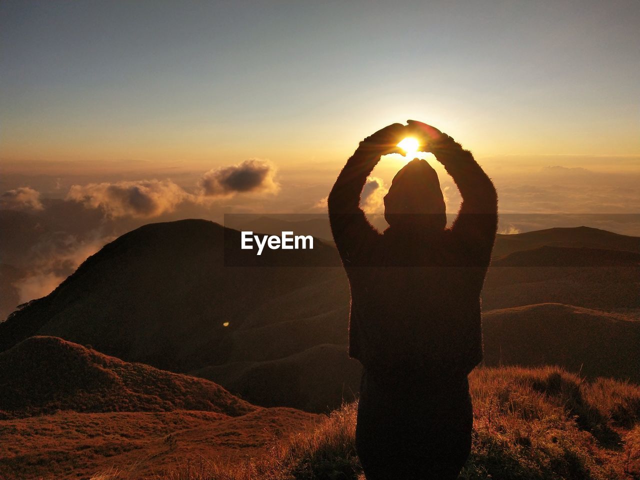 Rear view of woman with arms raised making hear shape while standing against sky during sunset