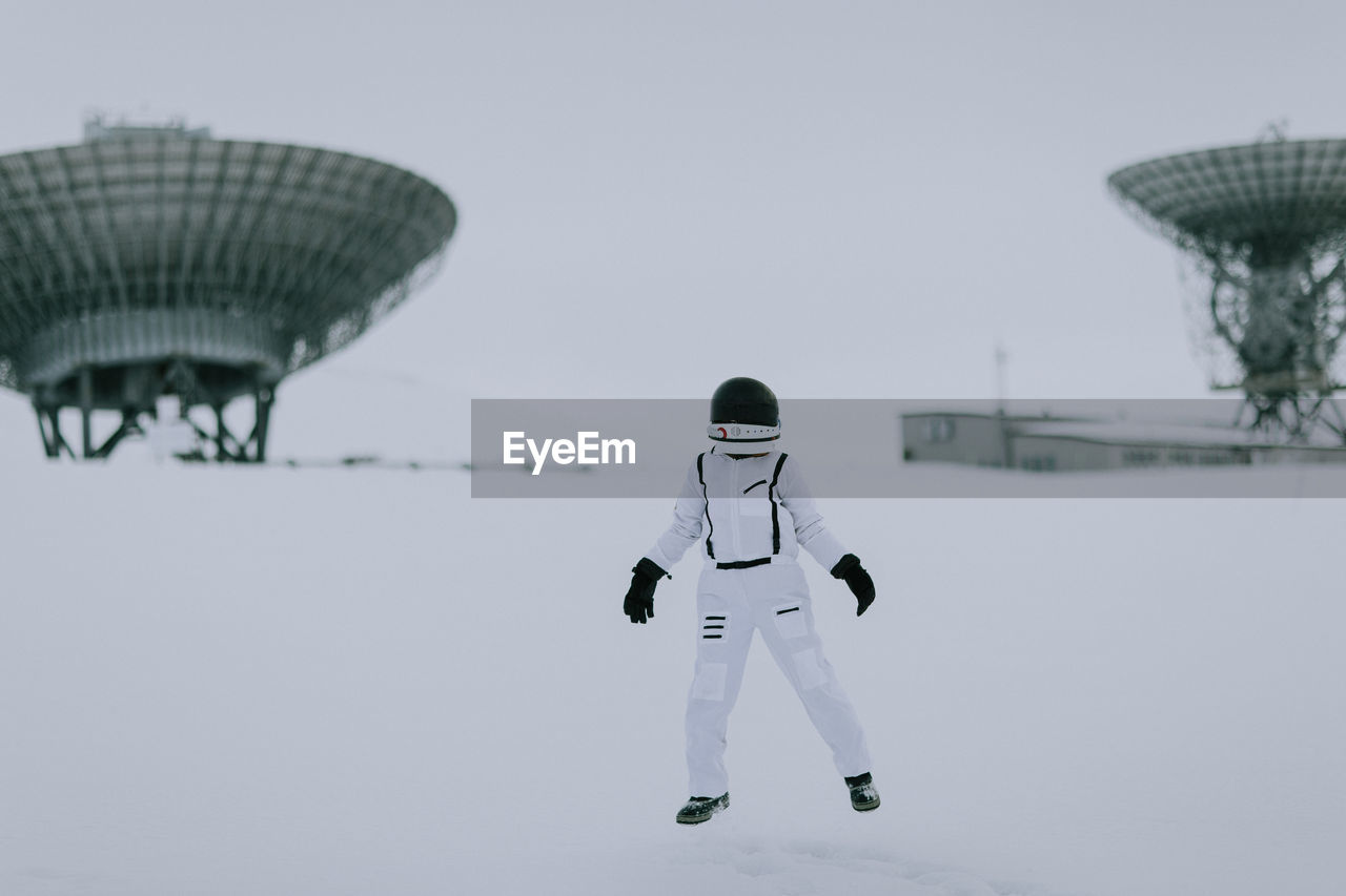 Unrecognizable cosmonaut in spacesuit standing in snowy valley in winter on background of huge radar antennas in svalbard