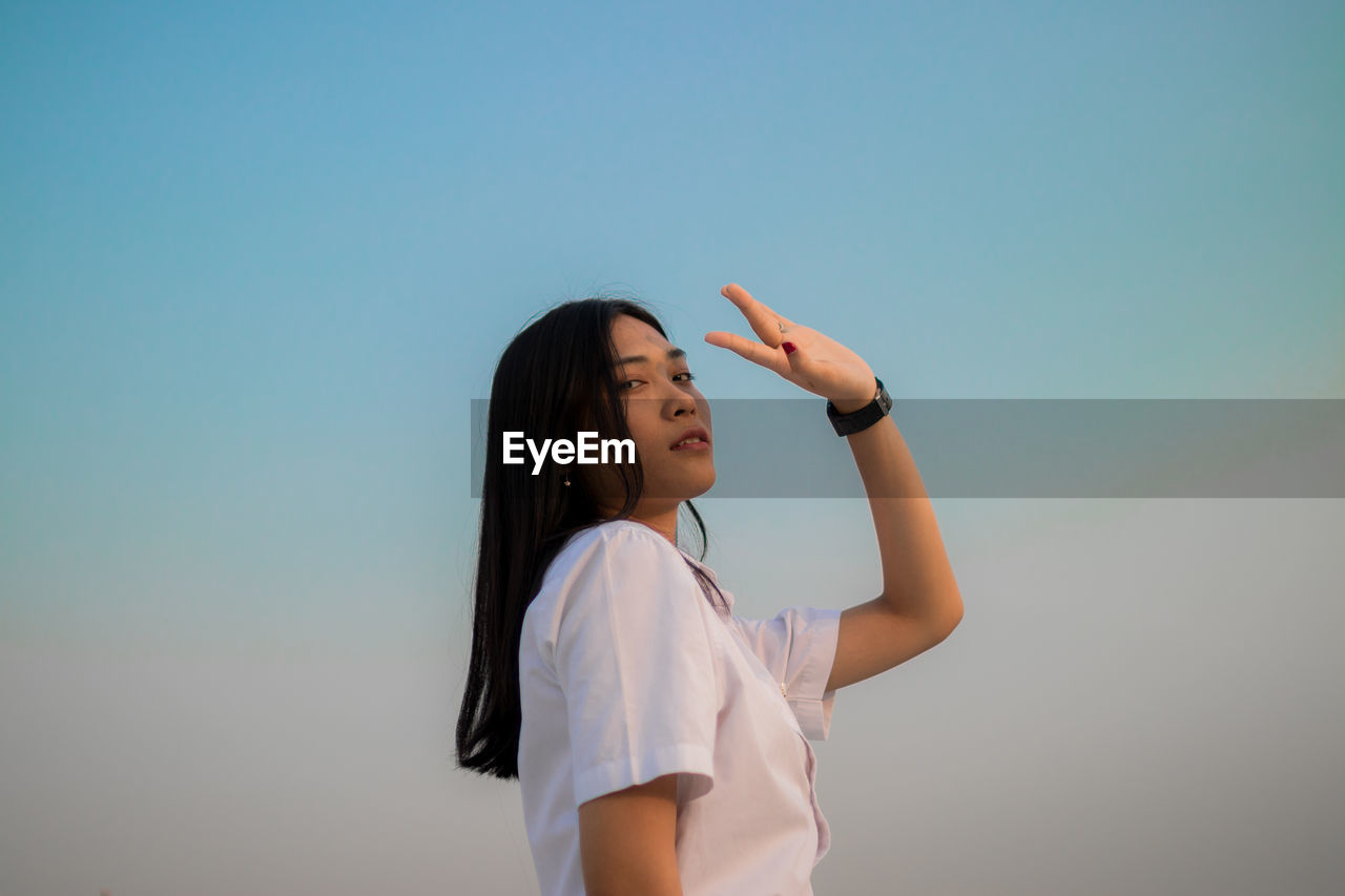 Low angle view portrait of young woman shielding eyes standing against sky during sunset