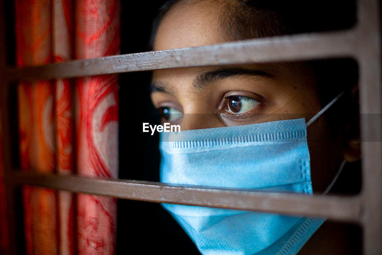 A bored asian young girl wearing a protection surgical face mask at looking through a window