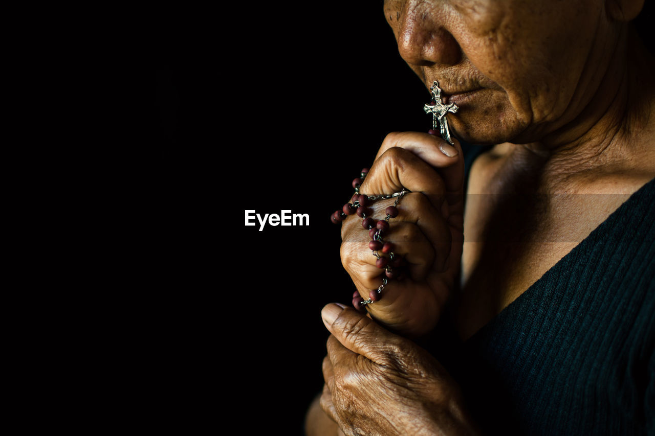 Close-up of senior woman holding rosary while praying against black background