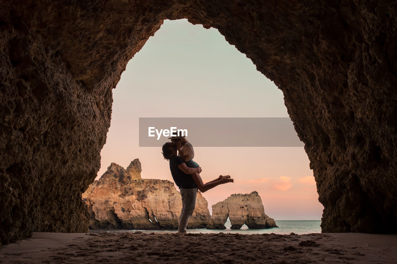 Full body side view unrecognizable man lifting and kissing girlfriend while standing in entrance of cave near sea in benagil caves in algarve, portugal
