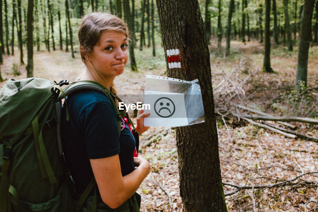 Portrait of woman showing paper with anthropomorphic face on tree trunk in forest