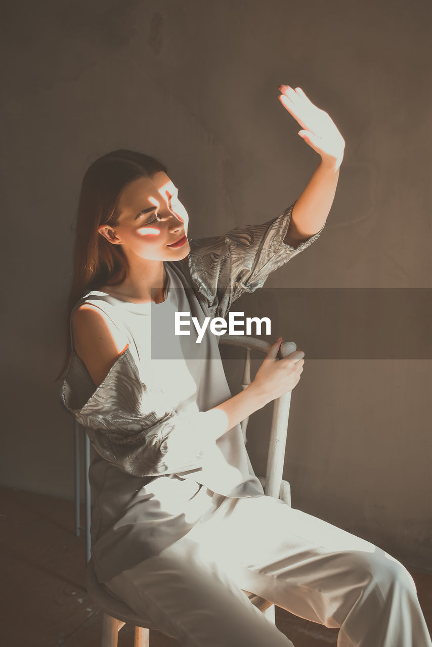 Young woman shielding eyes while sitting on chair against wall