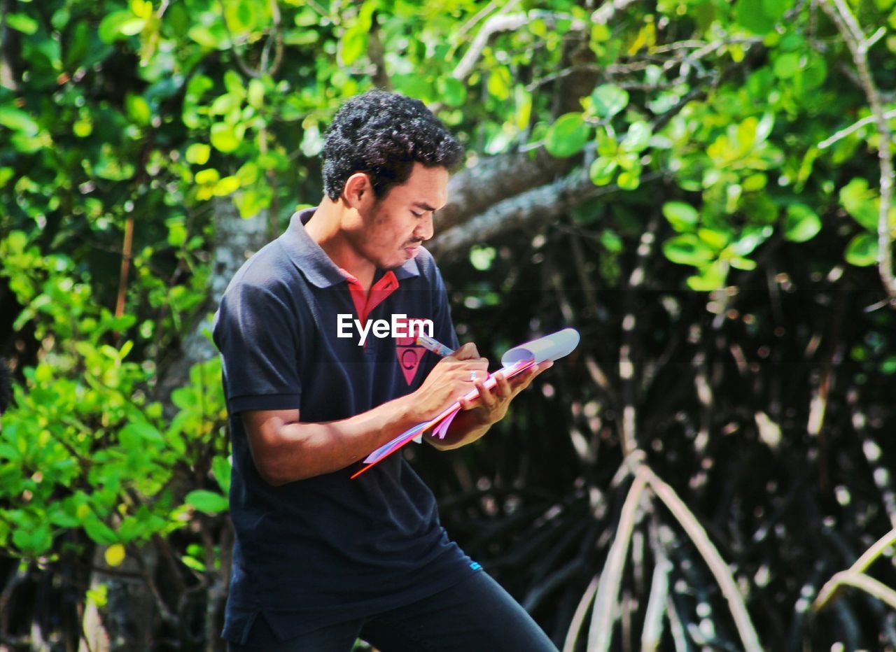 Young man writing on clipboard while standing against tree