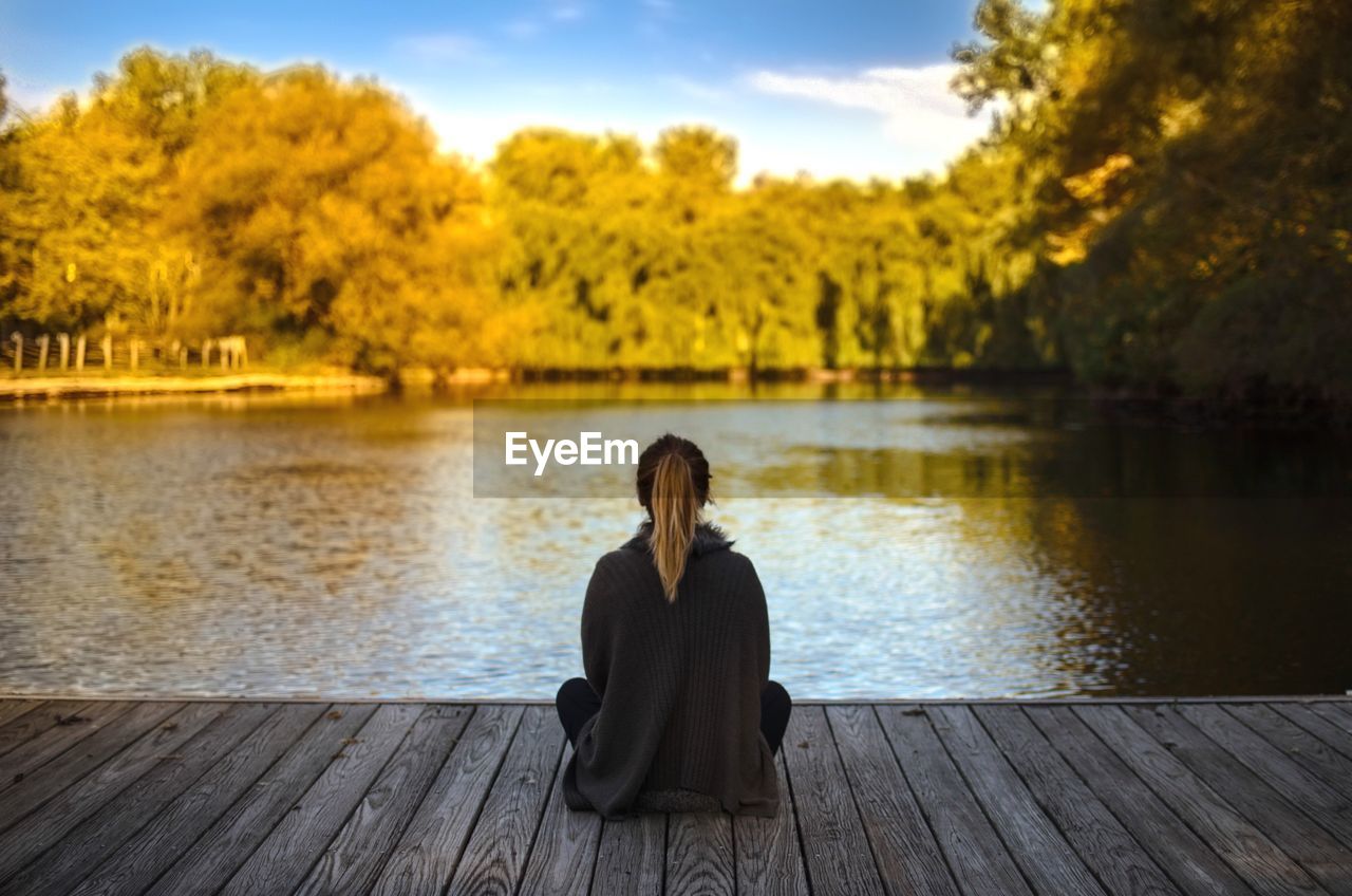 Young woman sitting on jetty by lake against sky