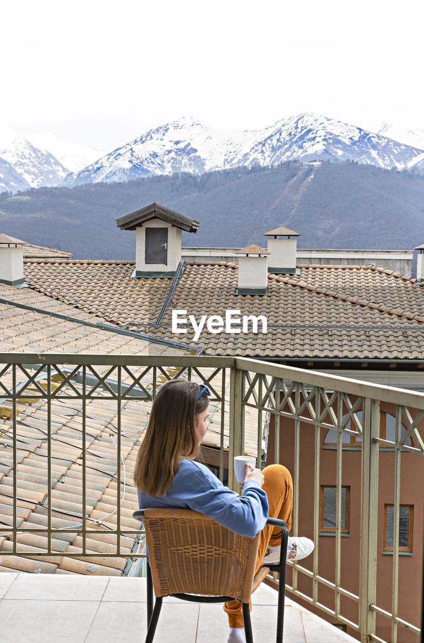 Rear view of young woman in blue sitting with mug of drink on terrace and looking at mountain view 