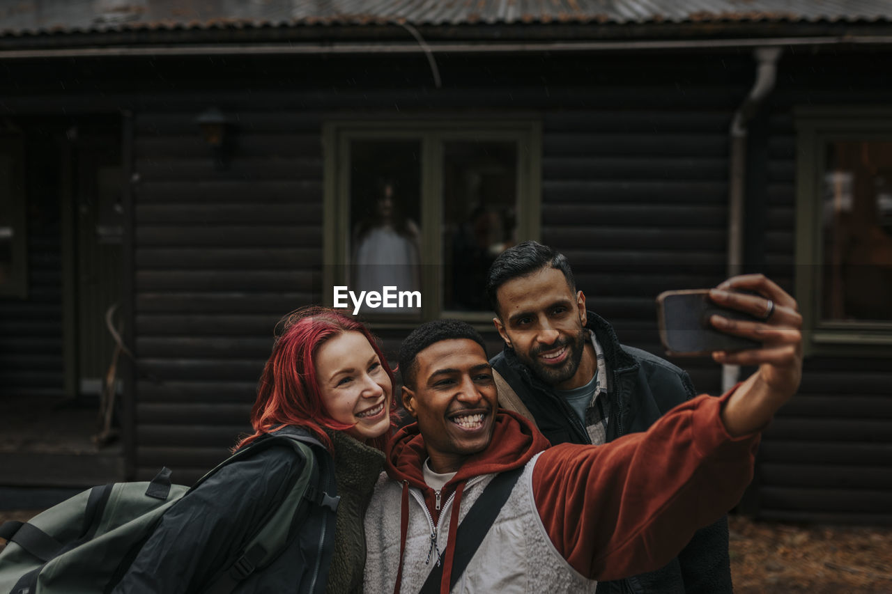 Happy multiracial friends taking selfie with smart phone while ghost at window in background