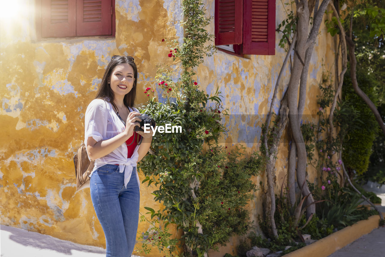 Portrait of smiling woman holding camera while standing outdoors