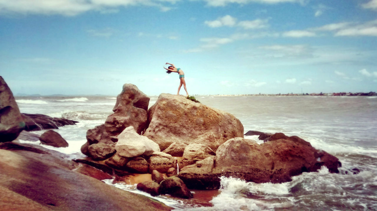 Mid distance view of young woman standing on rock at sea against blue sky