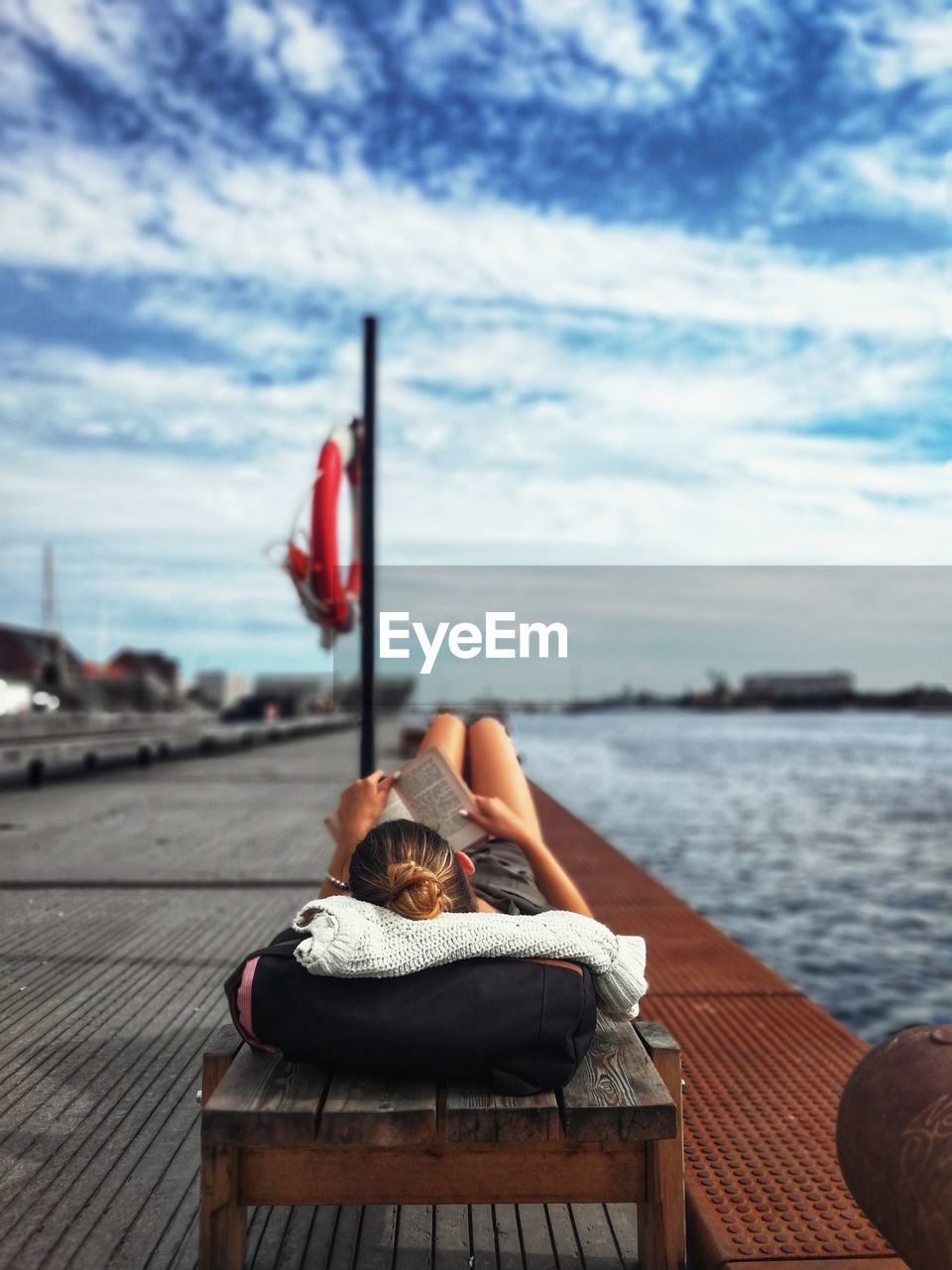 Woman reading book while lying at harbor against sky