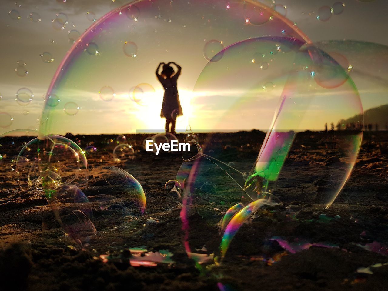 Silhouette girl seen through bubbles standing on sand against sky