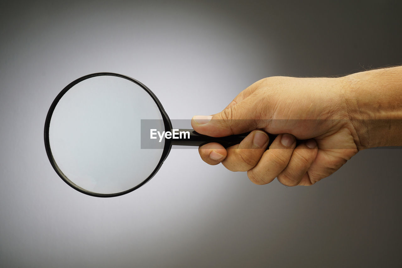 Close-up of hand holding magnifying glass against gray background