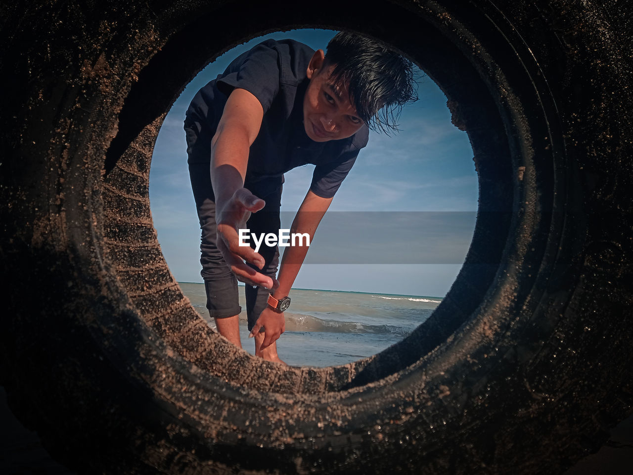 Young man gesturing while looking through tire against sky