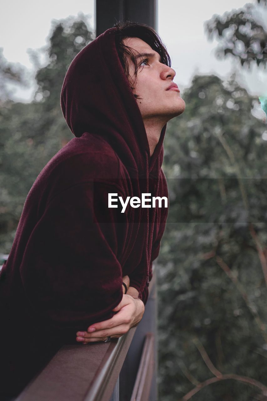 Side view of young man looking up standing by tree