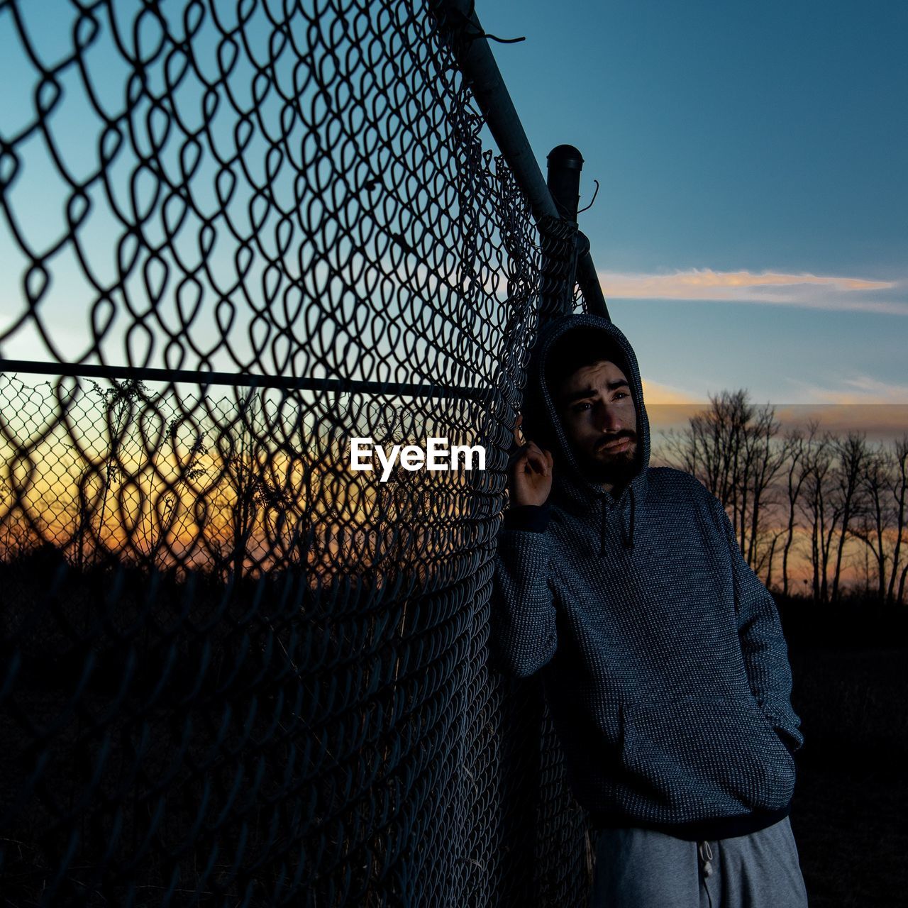 Thoughtful young man standing by fence against sky during sunset