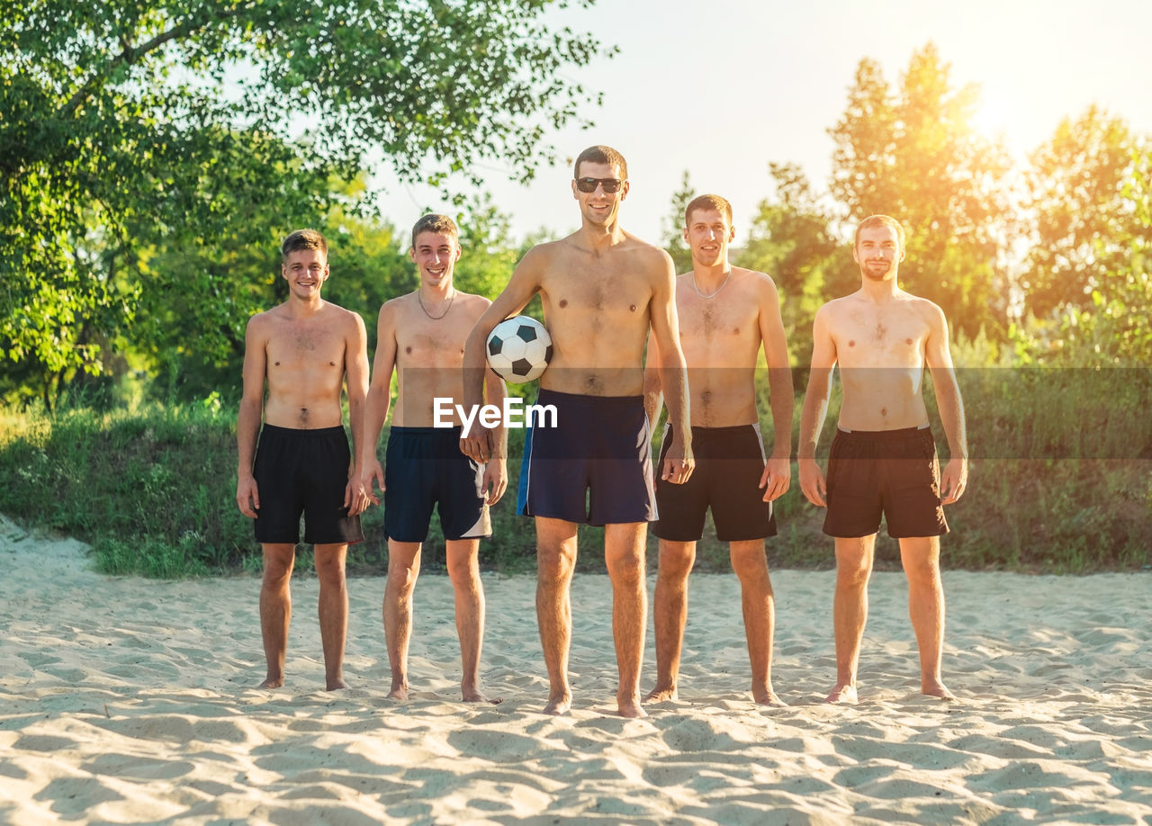 Group of happy beach soccer players stay with ball after football game on sand in summer sunny day.