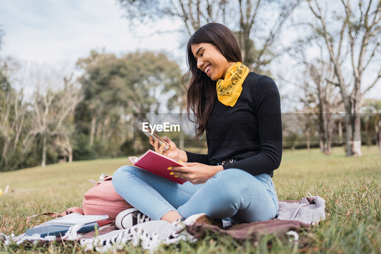 Young ethnic female university student in denim outfit browsing on smartphone while reading book in park