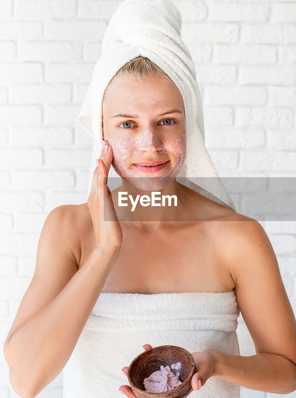 Woman wearing white bath towels on head doing spa procedures applying facial scrub