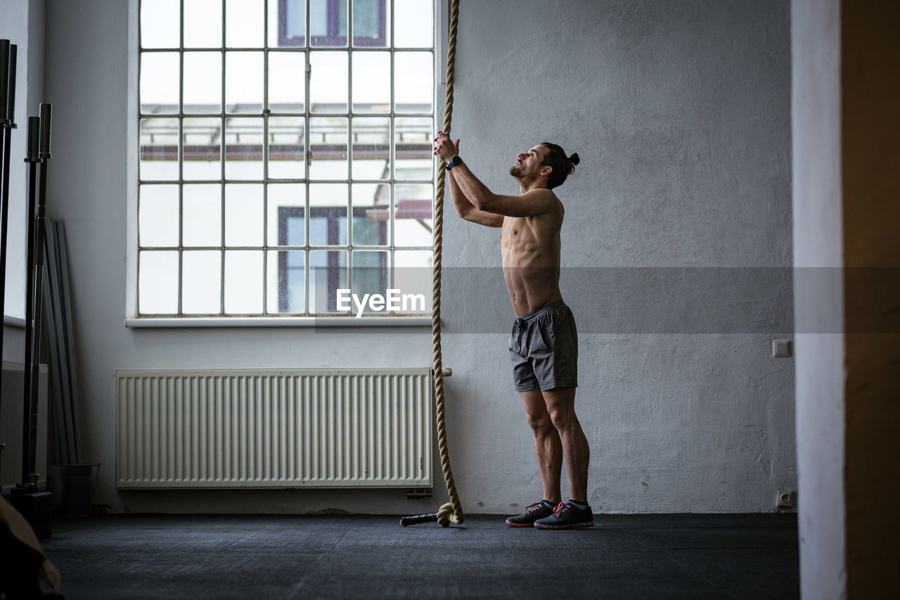 Full length of shirtless man standing against window