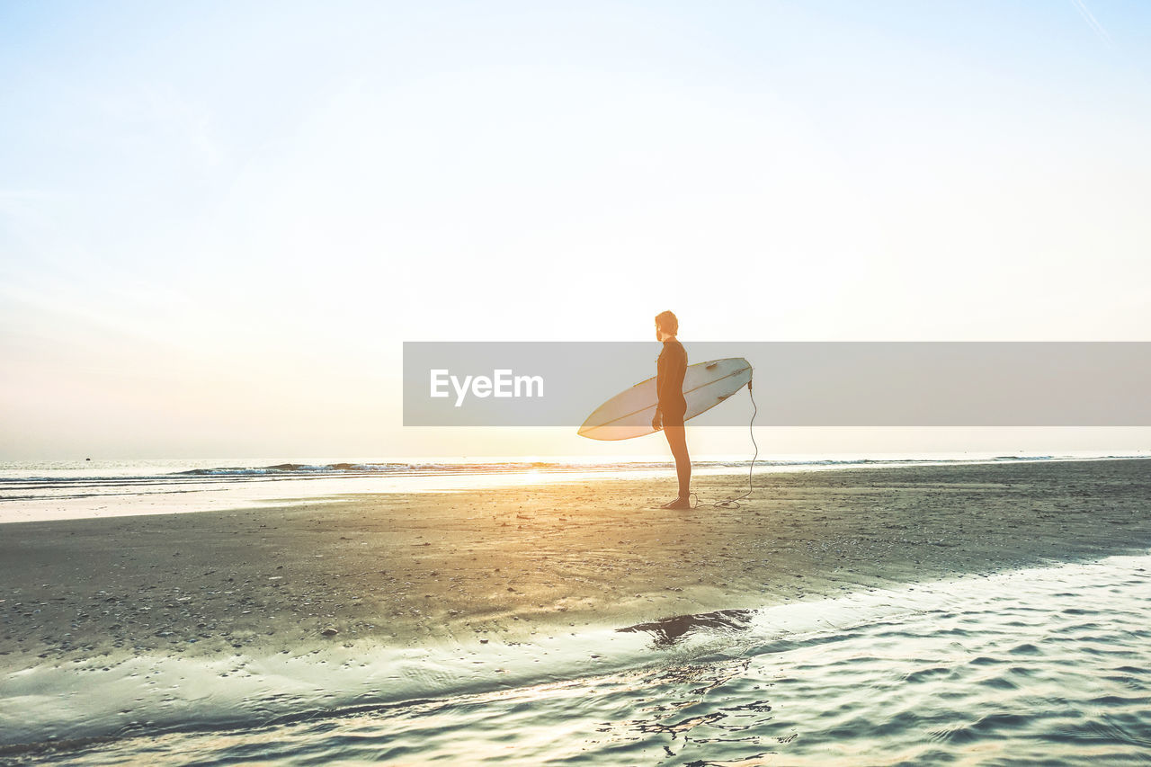 Full length of man with surfboard standing on beach against sky