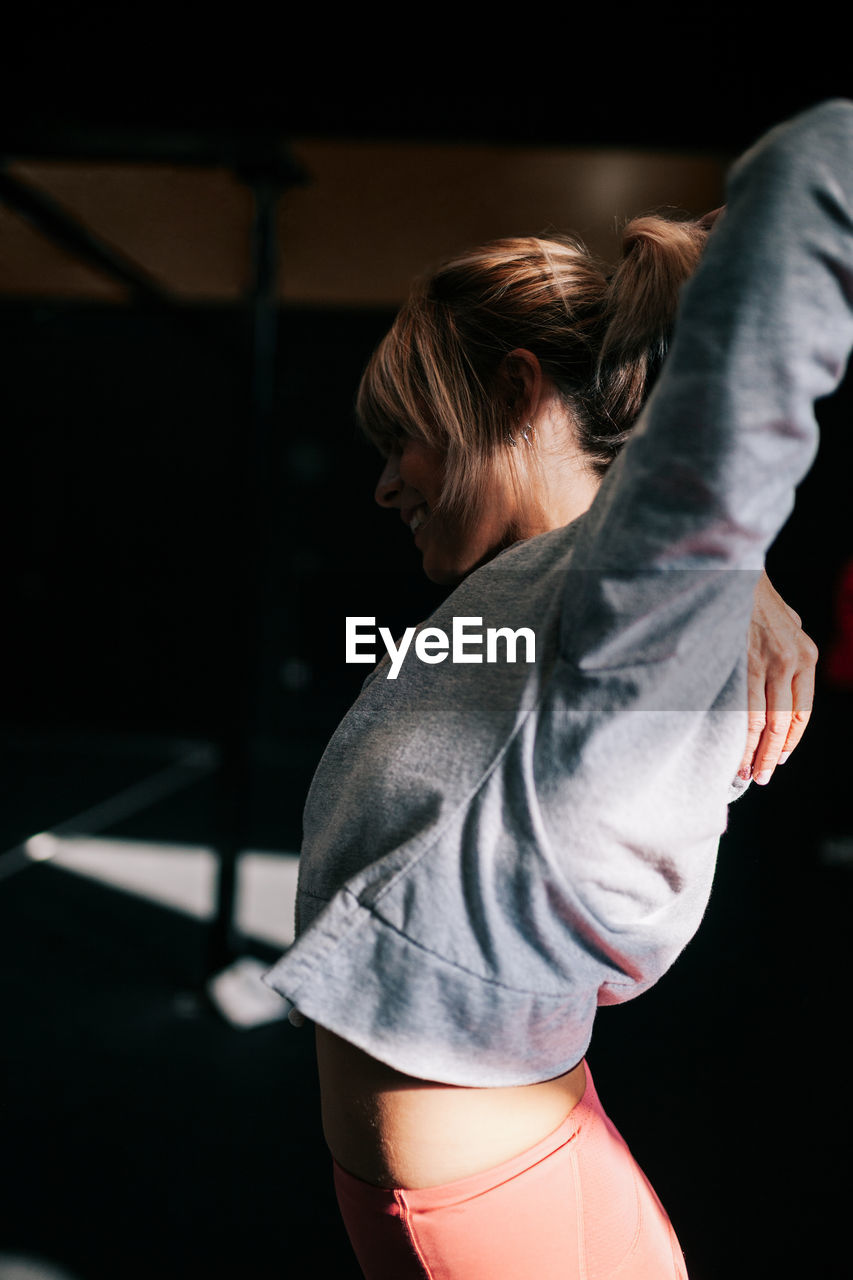 Side view of positive young fit female athlete in sportswear performing stretching exercise during workout in dark gym