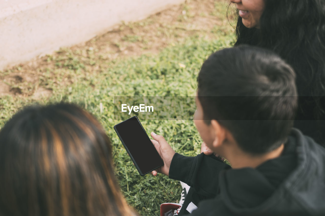 View from the back group of laughing latinos sitting on the ground in a park with a smartphone