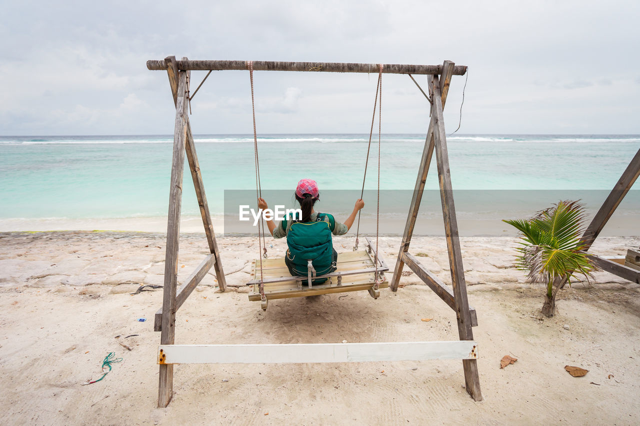 Back view of unrecognizable female traveler sitting on swings and enjoying fresh breeze while resting during summer holidays on sandy beach of hulumale island in maldives