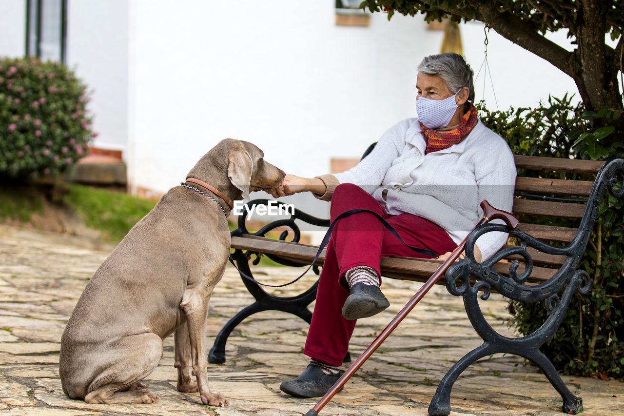 Full length of senior wearing mask with dog sitting outdoors