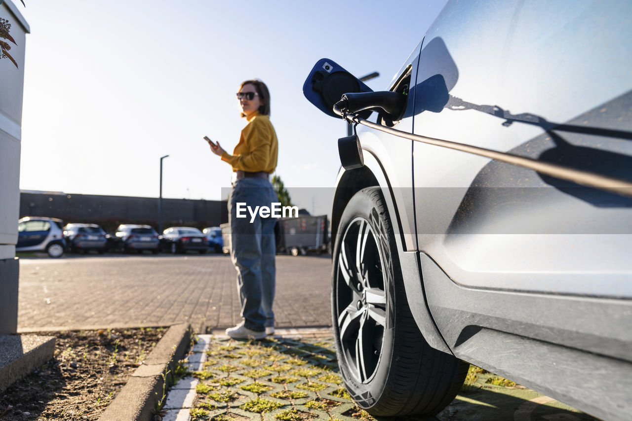 Electric car charging at station with woman in background holding smart phone on sunny day